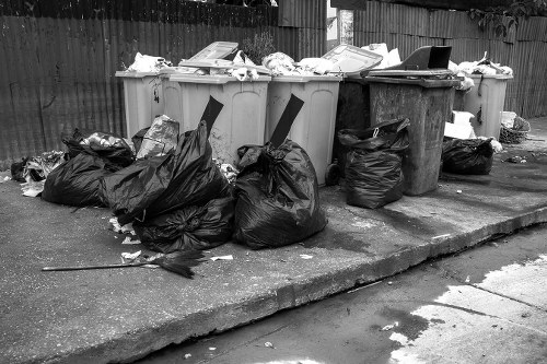 A row of five large wheelie bins is situated on a concrete driveway, with three beige bins at the front and two darker bins behind them. The beige bins are filled with various types of waste, some overflowing with papers and plastics visible at the top, while the darker bins appear to contain less visible material. Several black rubbish bags are piled on the left side of the bins, some partially torn open, with loose debris scattered around them on the pavement. The surrounding environment includes a wooden fence in the background and parts of a residential garden on the right, with the scene captured during daylight hours. The overall setting indicates a typical rubbish disposal area, suggesting services such as commercial waste removal in Marylebone might be required for proper collection and waste management from such a site.