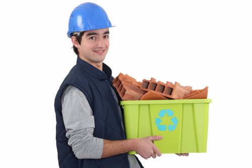 A young male waste collection worker wearing a blue safety helmet, a navy blue vest over a grey long-sleeved shirt, is holding a light green recycling bin with a blue recycling symbol on the side. The bin contains red clay building bricks stacked neatly. The background is plain white, emphasizing the worker and the bin. This image visually relates to rubbish removal services, showcasing a professional handling waste materials, with attention to safety gear and the recycling aspect of waste management, suitable for a website focused on commercial waste removal in Marylebone, London.