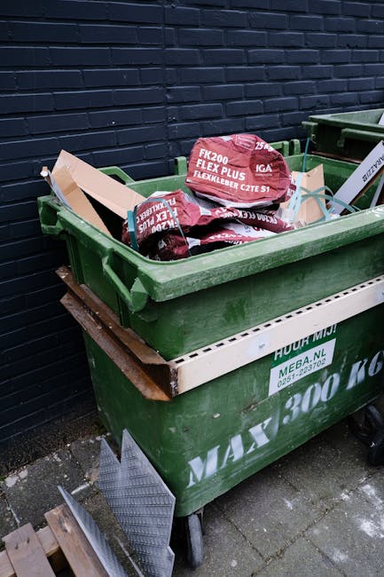 A narrow urban alleyway filled with a large, wheeled collection trolley in the foreground, covered in a weathered, grey fabric and positioned centrally on the paved surface. Behind it, there are stack